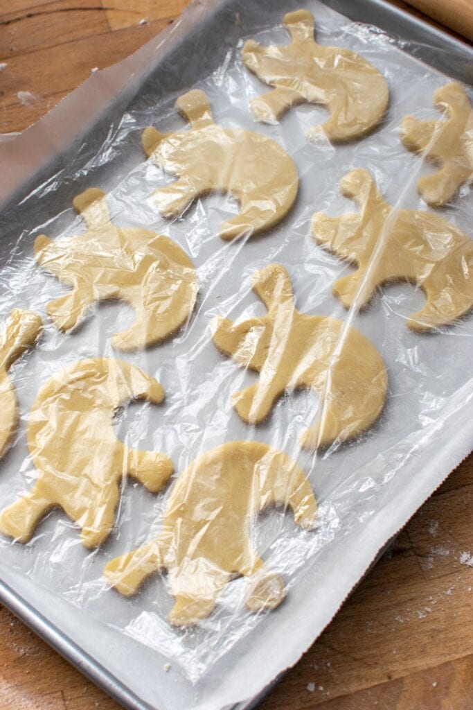 Halloween ghost cookie cut outs on a baking tray covered in plastic wrap to be chilled in the refrigerator.