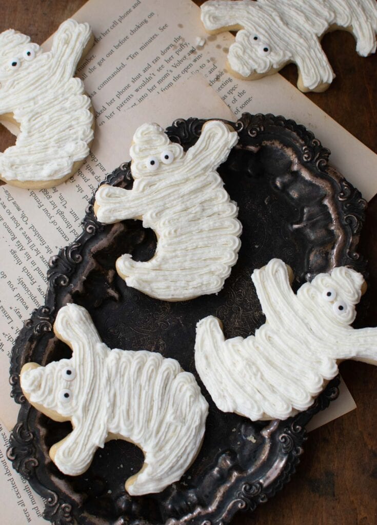 Halloween Ghost Cookies decorated with white frosting and candy eyes on a tarnished silver plate.