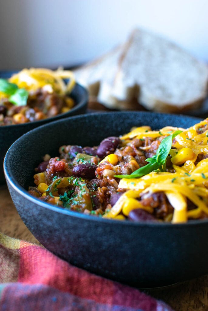 A close up of a black stoneware bowl filled with bean and lentil stew, topped with shredded cheddar cheese.