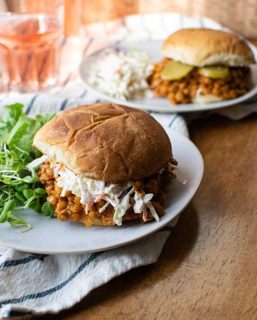Two lentil sloppy Joe's with a side of salad.  One Sloppy Joe it topped with Cole Slaw and the other is topped with Dill Pickles.