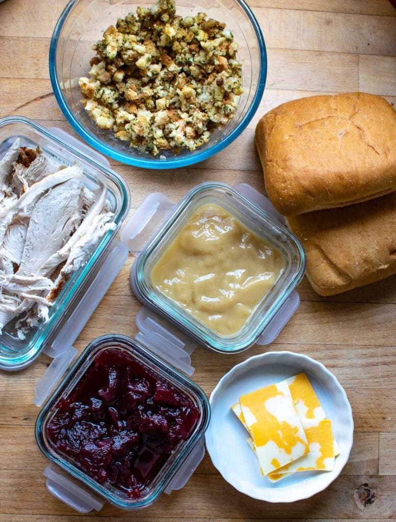 open containers of leftover turkey, stuffing, gravy and cranberry sauce with slices of cheese and rolls.