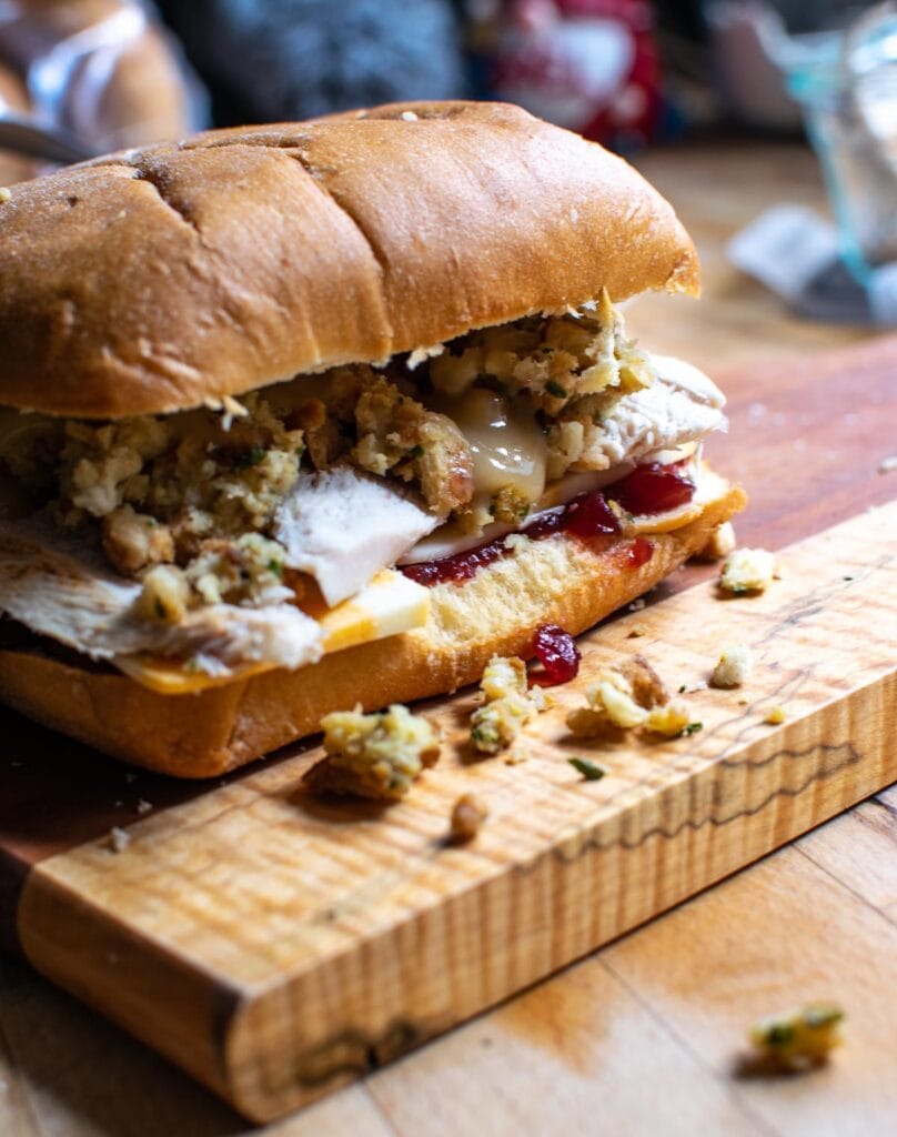 A loaded leftover turkey sandwich with layers of cranberry sauce, slice turkey, cheese, stuffing and gravy on a panini bun before being heated.
