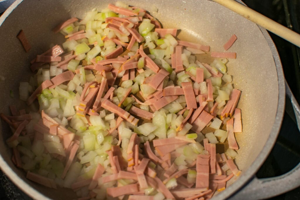 Sliced raw turkey bacon and chopped onions being browned in olive oil in a deep frying pan.