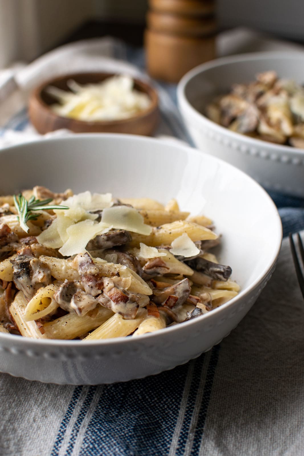 Two bowls of pasta coated with a cream mushroom and bacon sauce, topped with course salt, pepper and a sprinkle of parmesan cheese. Garnished with a sprig of rosemary.