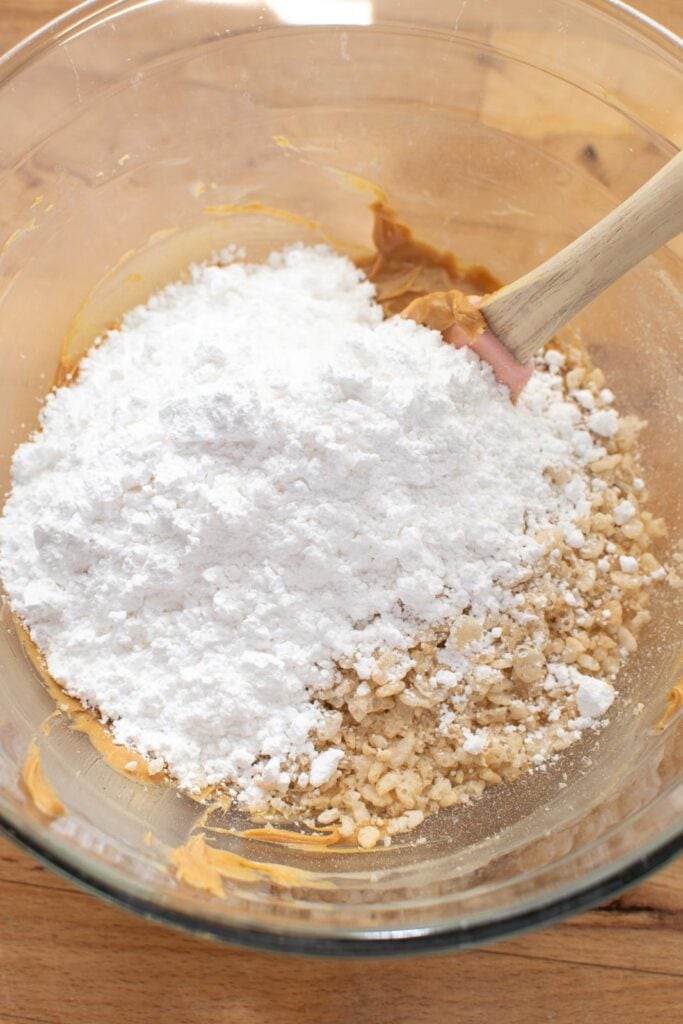 Rice Krispie cereal and confectioners sugar added to whipped peanut butter in a glass mixing bowl.