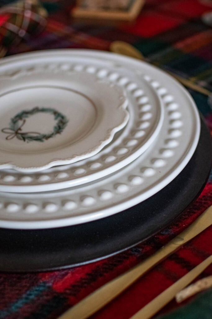 A place setting for Christmas table with a wood charger, white dinner and salad plates and a small dessert plate with a Christmas wreath.