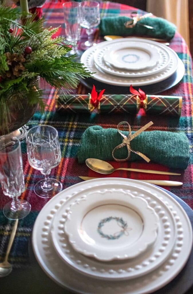 A dinner table set for Christmas with a red plaid tablecloth, white plates, tartan Christmas crackers and green linen napkins.