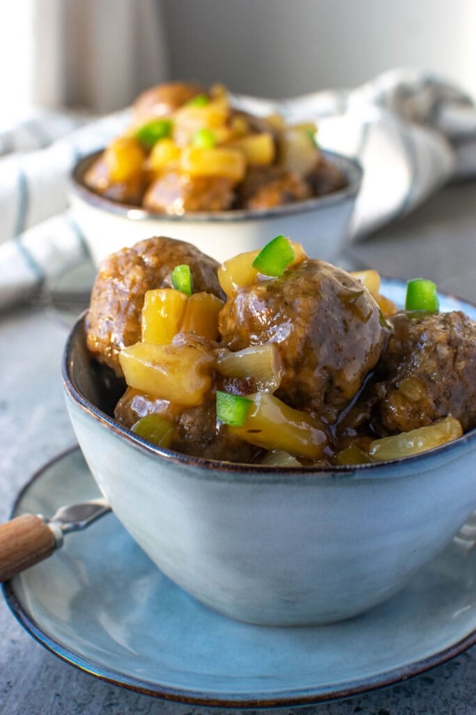 Two bowls of sweet and sour meatballs sitting on small plates and garnished with finely chopped green pepper.