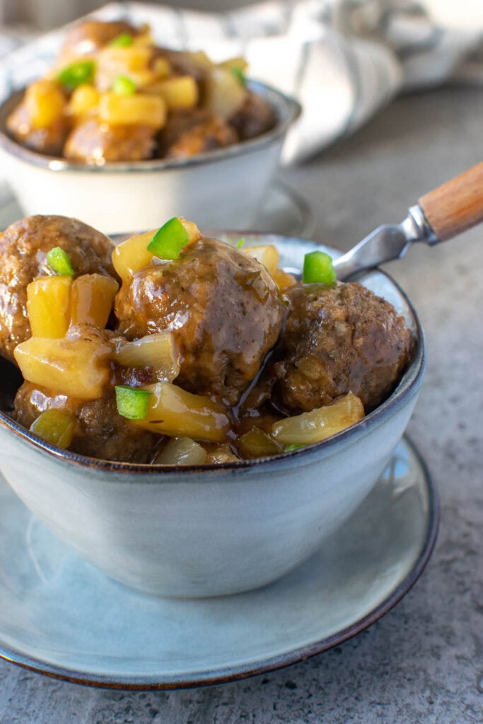 A bowl of sweet and sour meatballs garnished with finely chopped green pepper and a wooden handled fork in the back of the bowl.