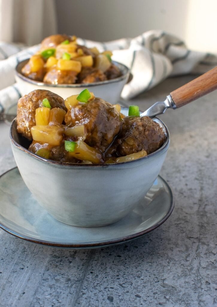 Two bowls of sweet and sour meatballs, one with a fork in the bowl and both garnished with tiny bits of chopped green pepper.