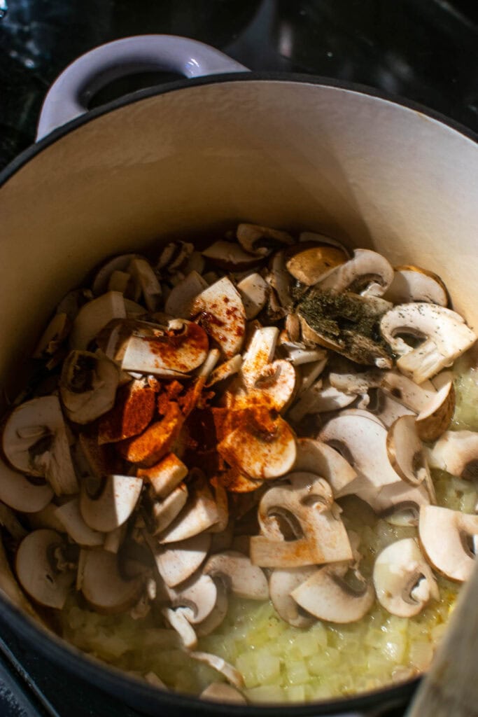 Sliced mushrooms added to a dutch oven pot with cooked onions, paprika, salt and pepper.