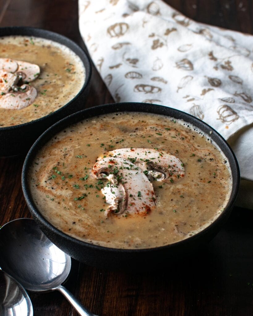 Two bowls of homemade cream of mushroom soup, with a napkin and two soup spoons.