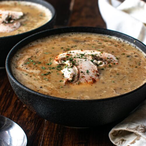 A large black stoneware bowl filled with cream of mushroom soup and garnished with fresh mushrooms.