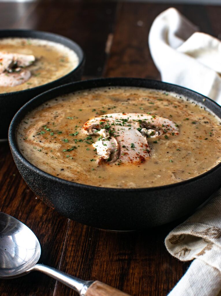 A large black stoneware bowl filled with cream of mushroom soup and garnished with fresh mushrooms.