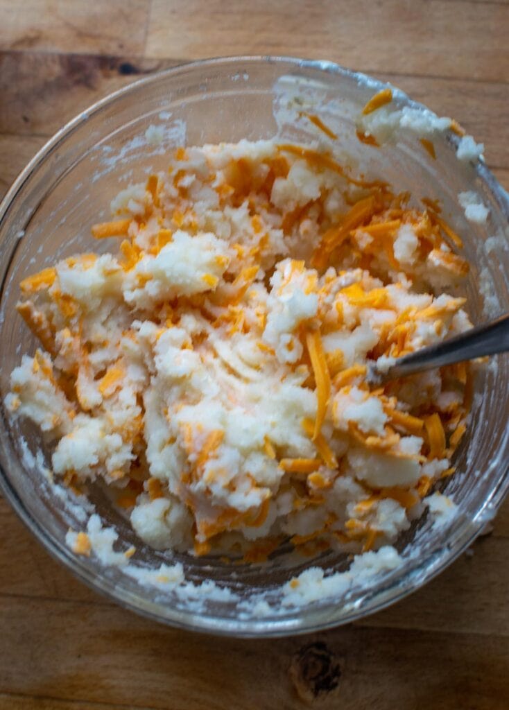 Mashed potatoes, shredded cheddar cheese and onion salt mixed together in a glass mixing bowl.