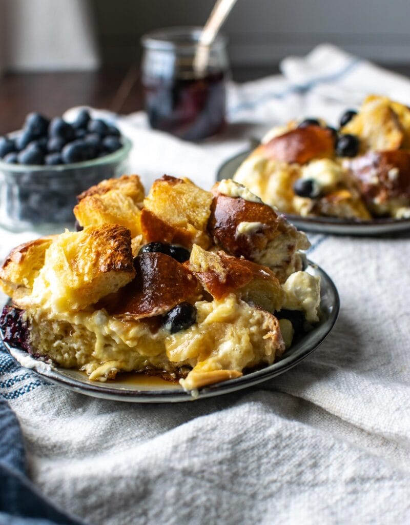 Two plates of blueberry cream cheese French toast casserole and a bowl of fresh blueberries to the side.