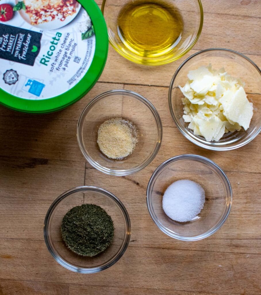 Ingredients to make a baked ricotta cheese dip including ricotta, parmesan, dill, salt and pepper, and olive oil.
