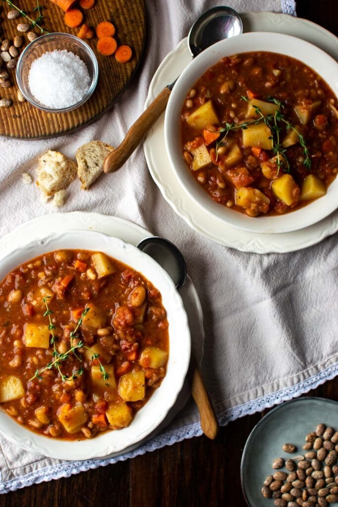 2 large bowls of bean and potato soup on top of a beige kitchen towel.  Wood handled spoons sit to the side of the bowls and small chunks of bread sit between the bowls.