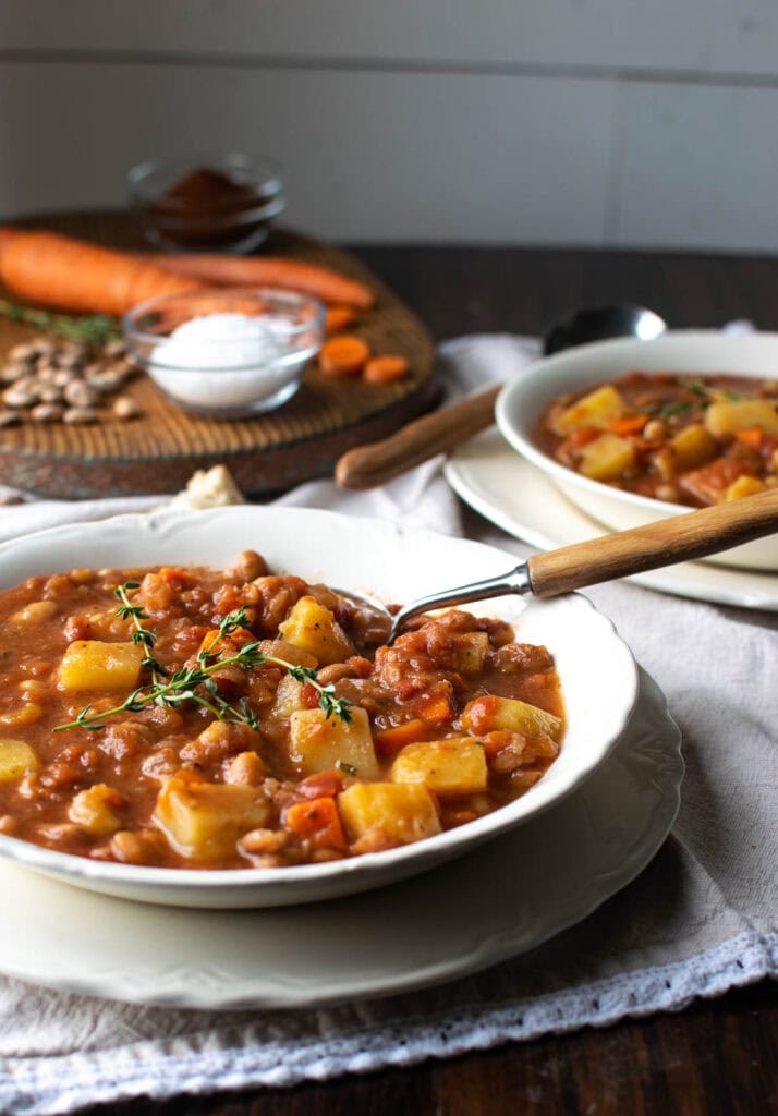 A bowl of bean soup with potatoes with a wood handled spoon in the bowl. In the background is another bowl of soup and a board with salt, beans and raw carrots chopped up.