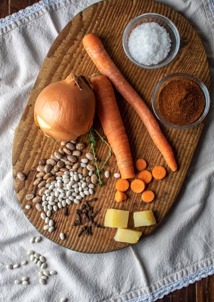 Some of the ingredients required to make potato bean soup, such as carrots, an onion, dried beans, chili powder, salt, cloves and chopped potatoes arranged on a cutting board.