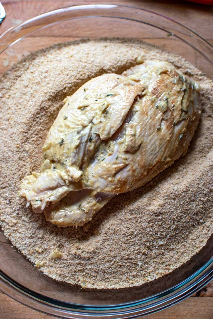 A mustard coated chicken breast in a plate of bread crumbs.