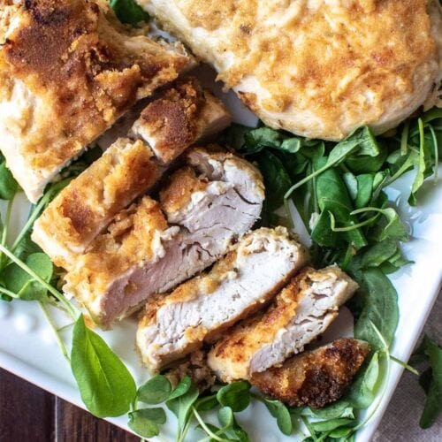 A crispy baked Dijon chicken breast set on top of greens on a platter with a few pieces sliced off to show the inside.