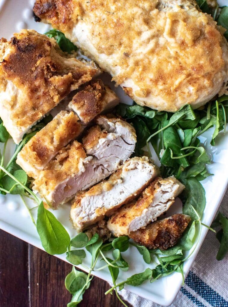 A crispy baked Dijon chicken breast set on top of greens on a platter with a few pieces sliced off to show the inside.