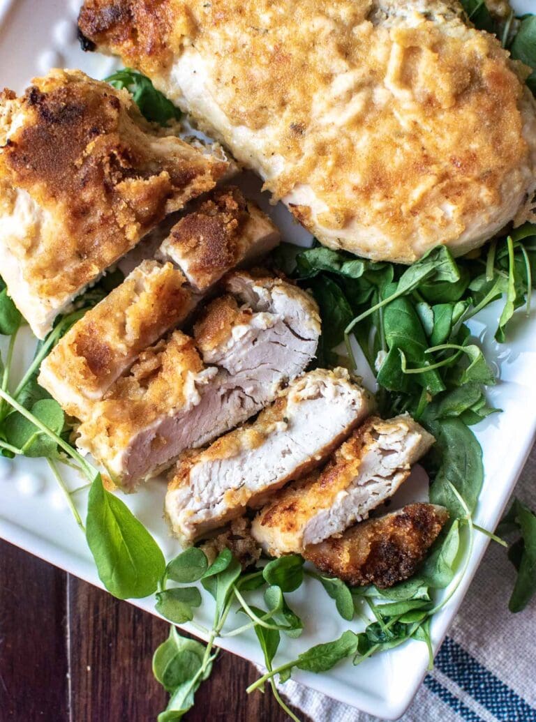 A crispy baked Dijon chicken breast set on top of greens on a platter with a few pieces sliced off to show the inside.