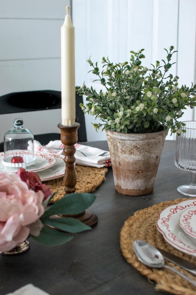 A faux green plant, white candle in wood candlesticks, faux pink roses all on a dinner table for Valentines Day.