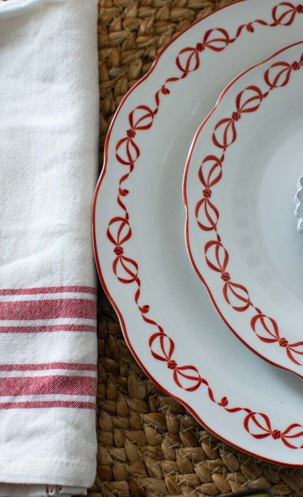 Dinner and salad plates with a bow design on the scalloped edges are beside a red and white napkin.
