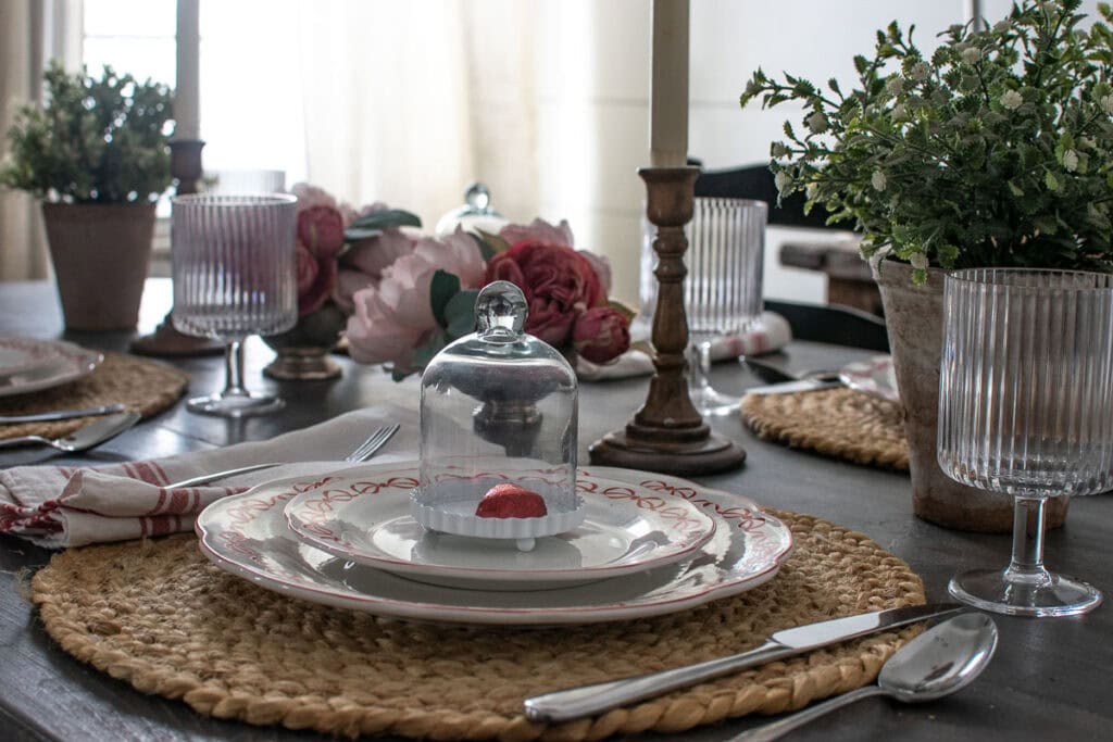 A heart shaped chocolate wrapped in red tin foil under a tiny cloche that is sitting on top of a dinner plate.