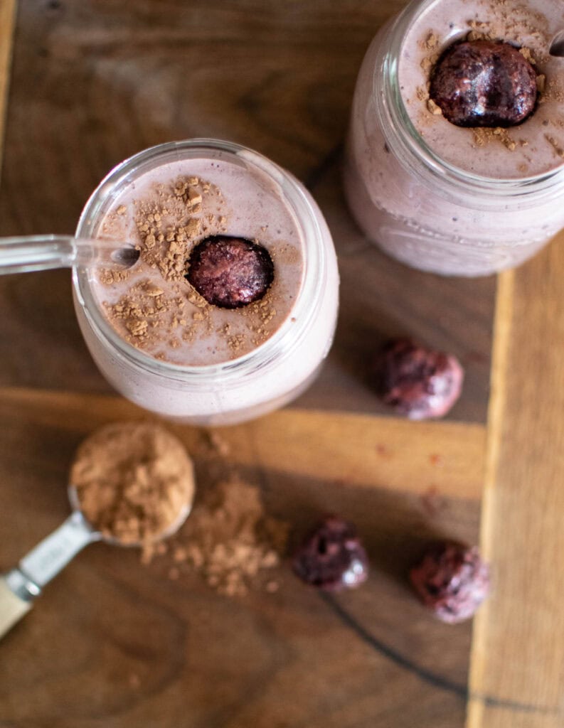 Overhead view of two mason jars filled with a chocolate smoothie, with a spoon of cocoa powder and a few frozen cherries beside them.