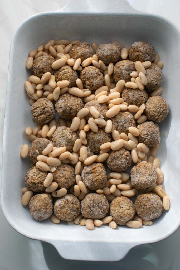 White kidney beans sprinkled over frozen meatballs in a glass casserole dish.