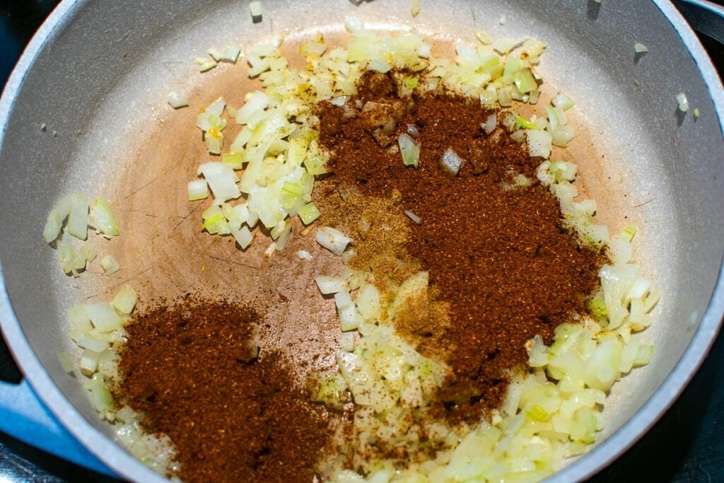 Chili powder and cumin sprinkled over chopped cooked onions in a frying pan on the stove top.