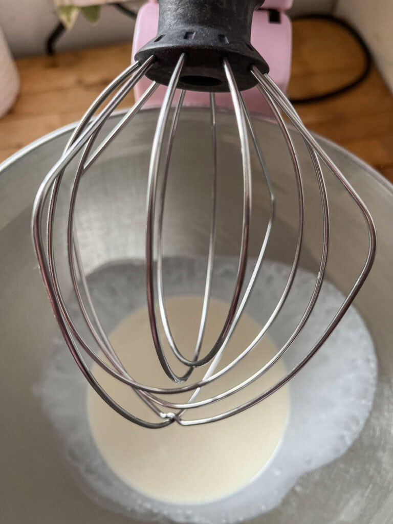 Pink Kitchen Aid stand mixer fitted with a whisk attachment and a mixing bowl with whipping cream.