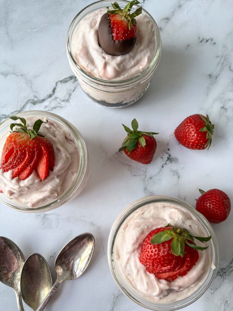 On overhead view of 3 jars of strawberry mousse with 3 fresh strawberries and 3 spoons to the side.