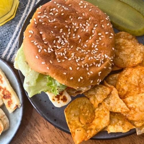 Grilled Halloumi Sandwich on a sesame seed bun and a side of potato chips and dill pickle slices.