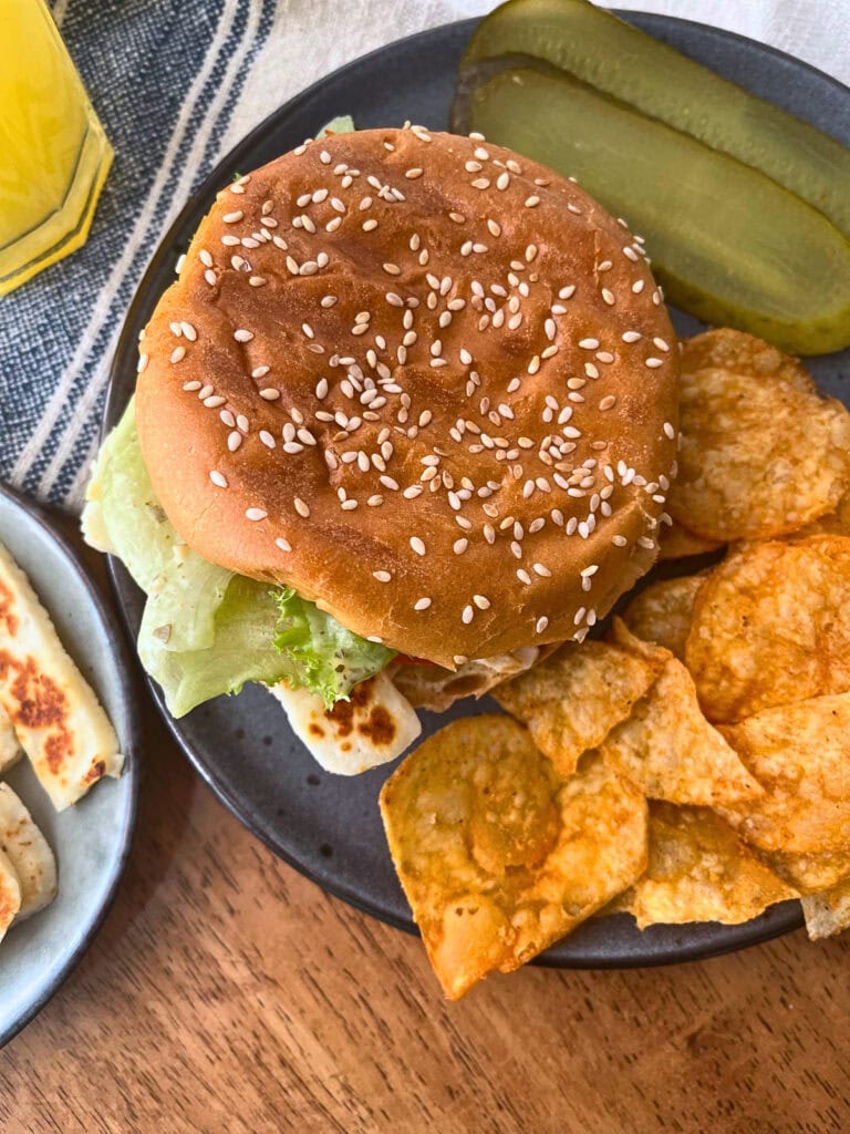 Grilled Halloumi Sandwich on a sesame seed bun and a side of potato chips and dill pickle slices.
