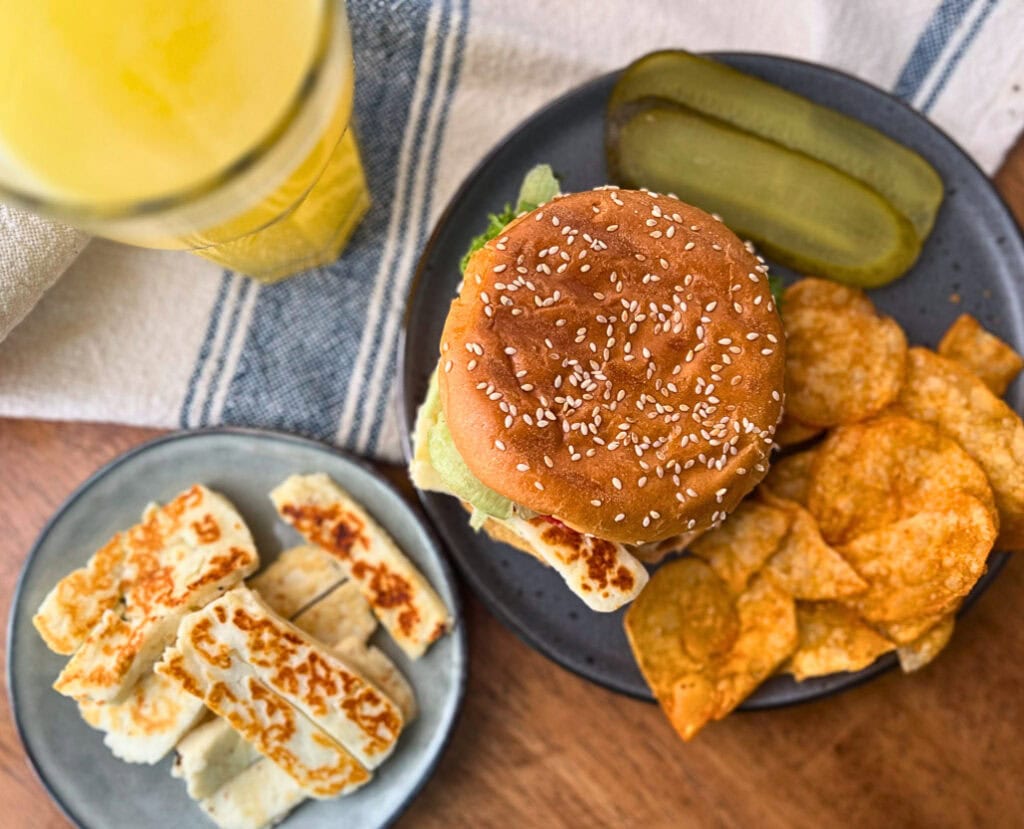 A plate of crispy halloumi cheese beside a sandwich made on a brioche bun with the halloumi cheese, tomato, lettuce and creamy pesto sauce, with a side of potato chips and sliced pickles.