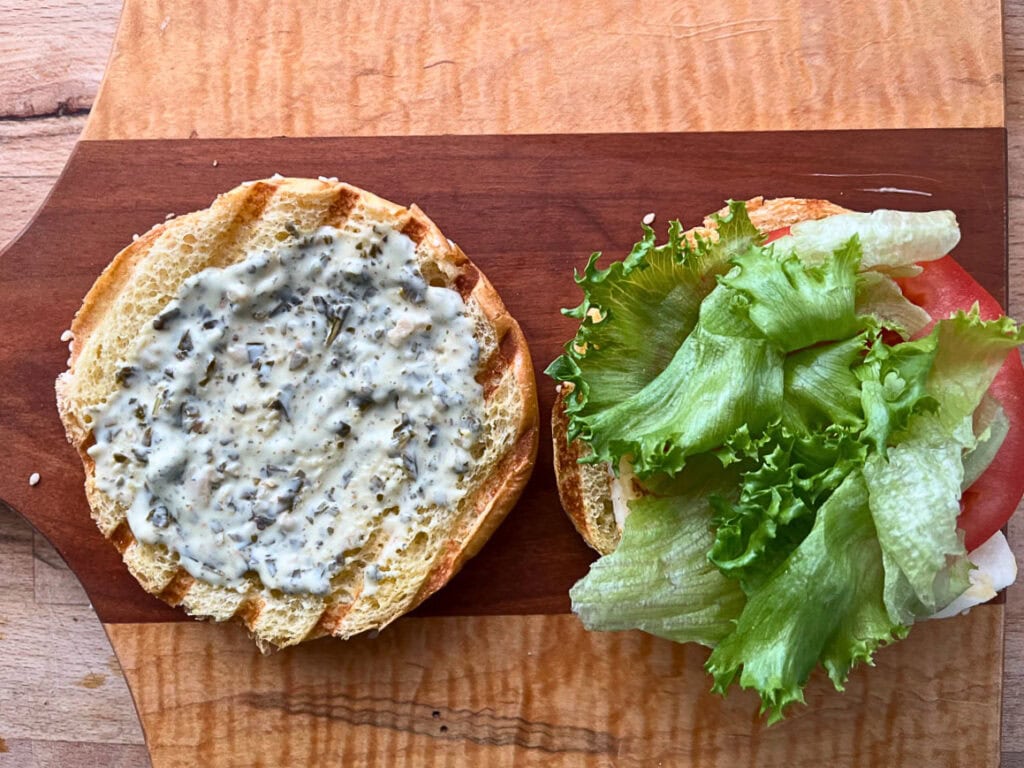 An open bun with the bottom one topped with a creamy pesto sauce, halloumi cheese, sliced tomato, and lettuce. The top side of the bun is topped with creamy pesto sauce.