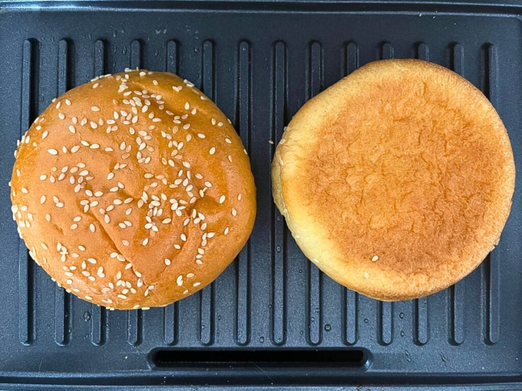 A sesame brioche bun toasting on a grill, cut side down.