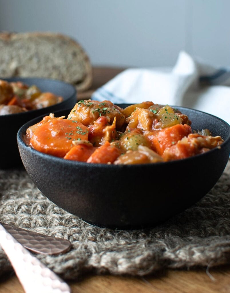A bowl of chicken and vegetable stew in a black ceramic bowl with a loaf of bread in the background.