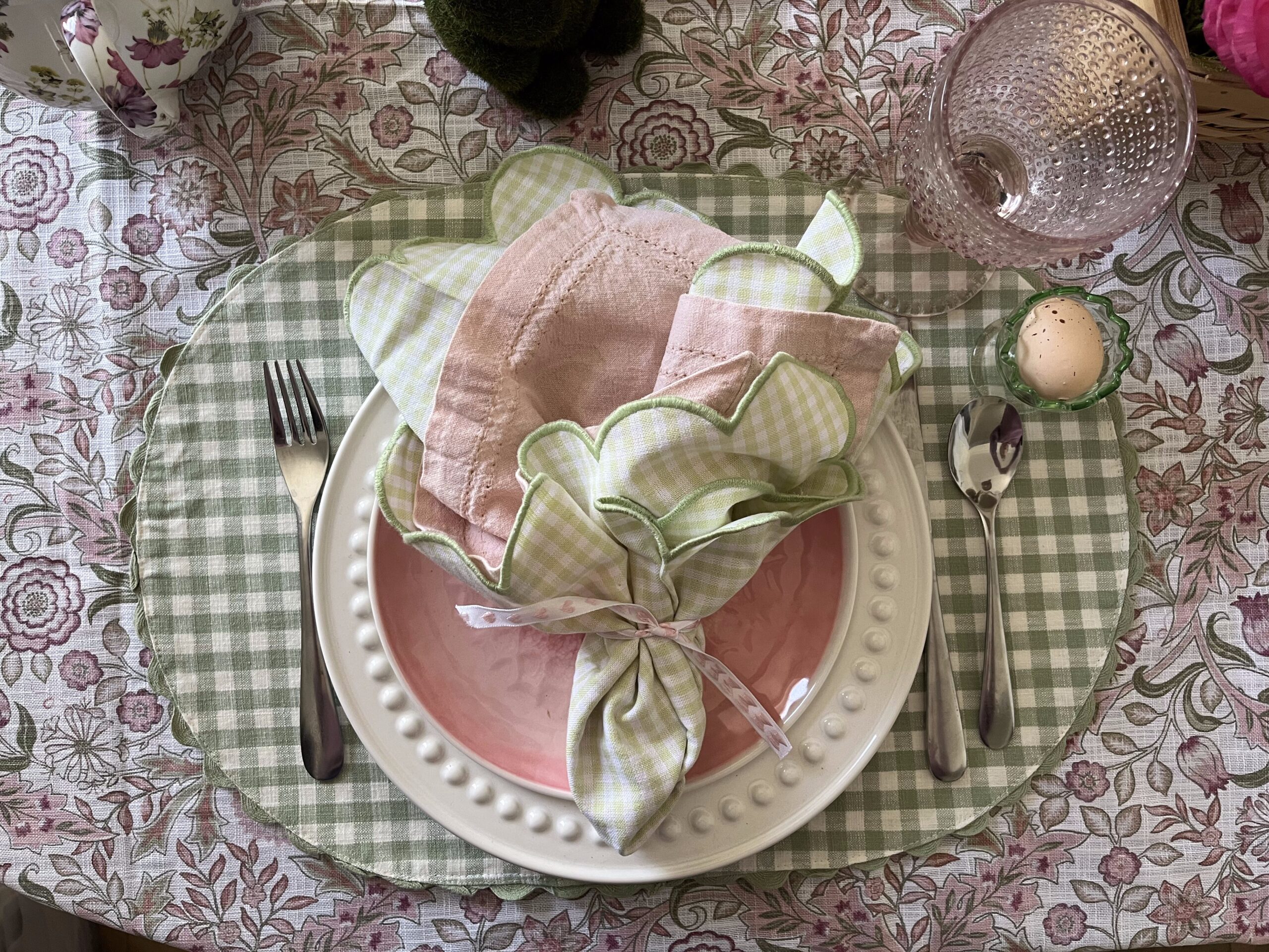 An Easter place setting featuring a green gingham place mat on top of a floral tablecloth, with white and pink plates.