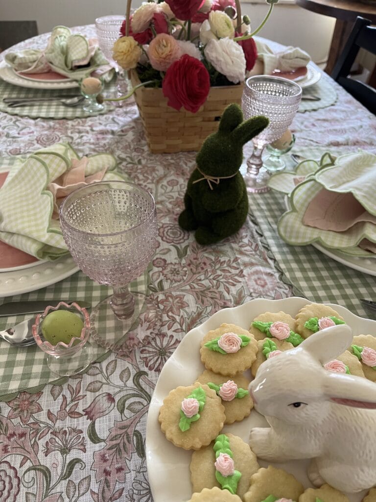 A plate of shortbread cookies decorated with green leaves and pink flowers sit on a cake plate with a ceramic bunny in the centre.