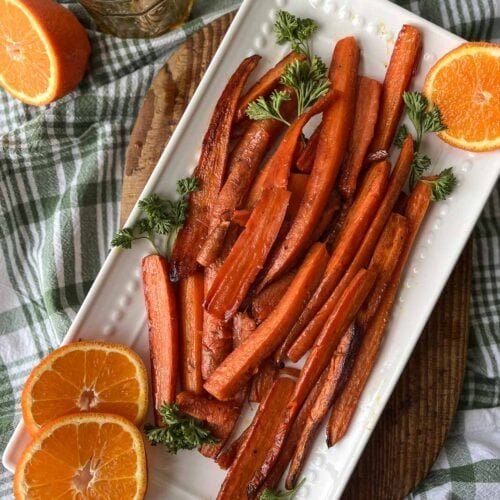 A white serving plate with roasted orange honey glazed carrots garnished with parsley and fresh orange slices.