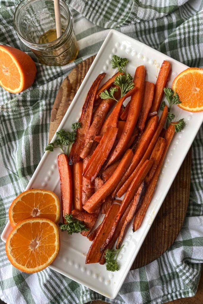 A white serving plate with roasted orange honey glazed carrots garnished with parsley and fresh orange slices.