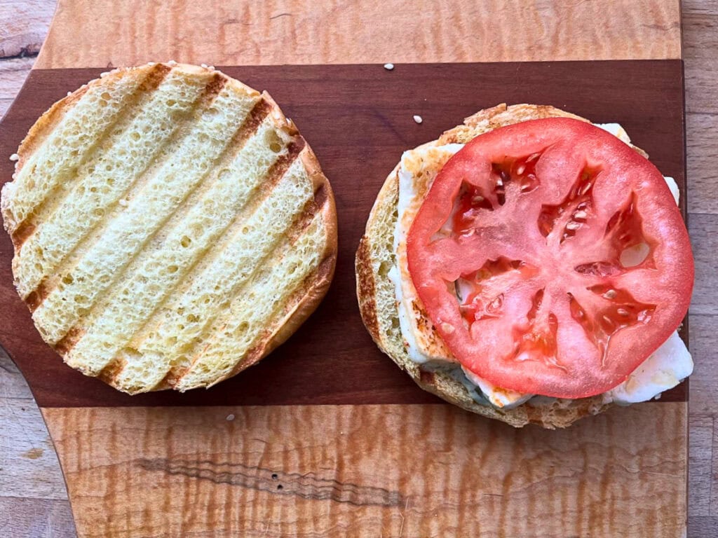 A halloumi cheese sandwich being assembled on a brioche bun, including tomato and creamy pesto sauce.