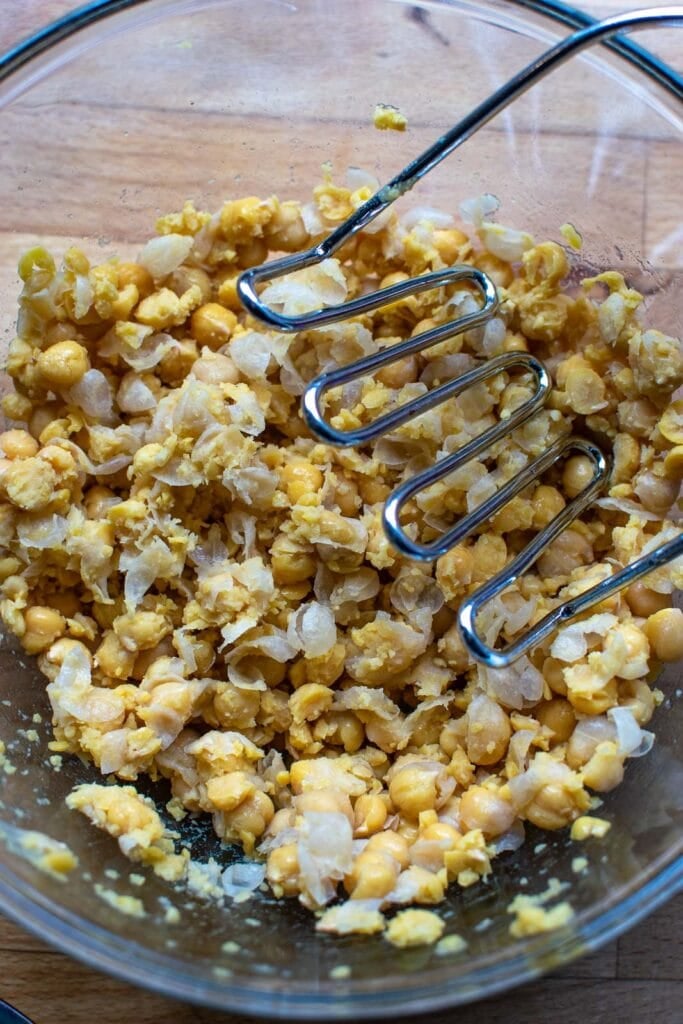 Drained chickpeas being mashed with a potato masher.