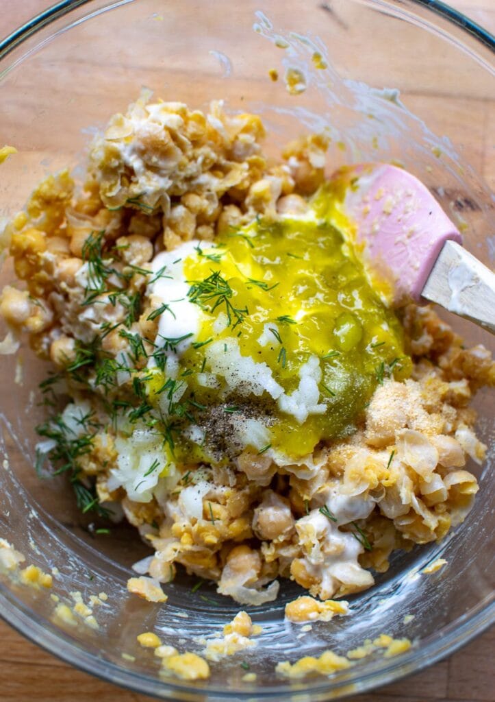 Mayonnaise, dill relish, chopped fresh dill, chopped onion and spices added to mashed chickpeas in a glass bowl.