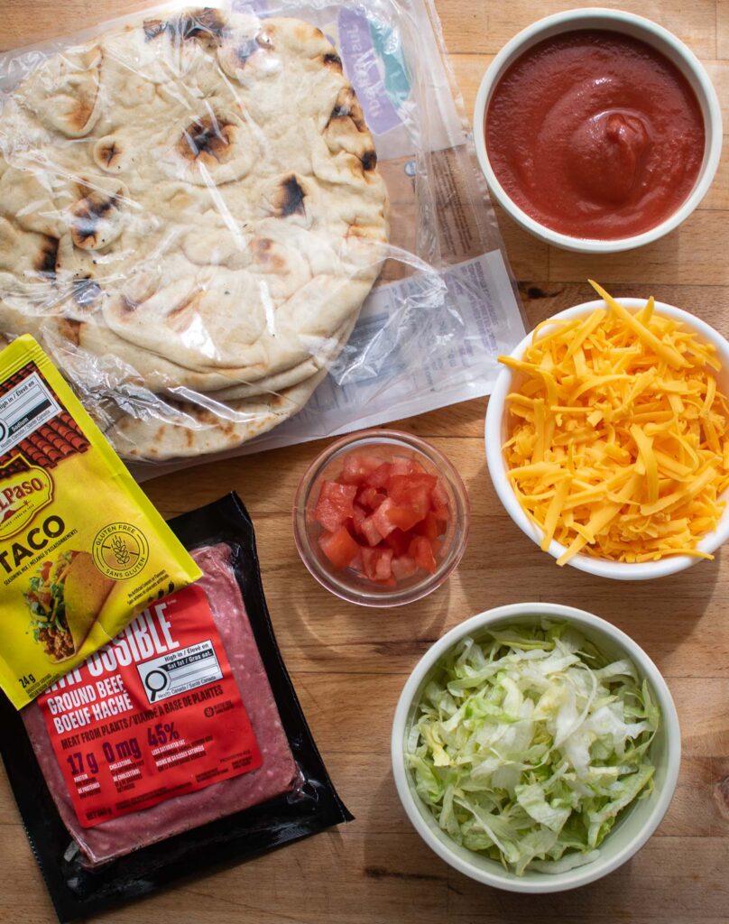 Ingredients to make taco pizza on Naan bread including shredded cheddar cheese, chopped lettuce and tomato, taco seasoning, ground beef and tomato sauce.